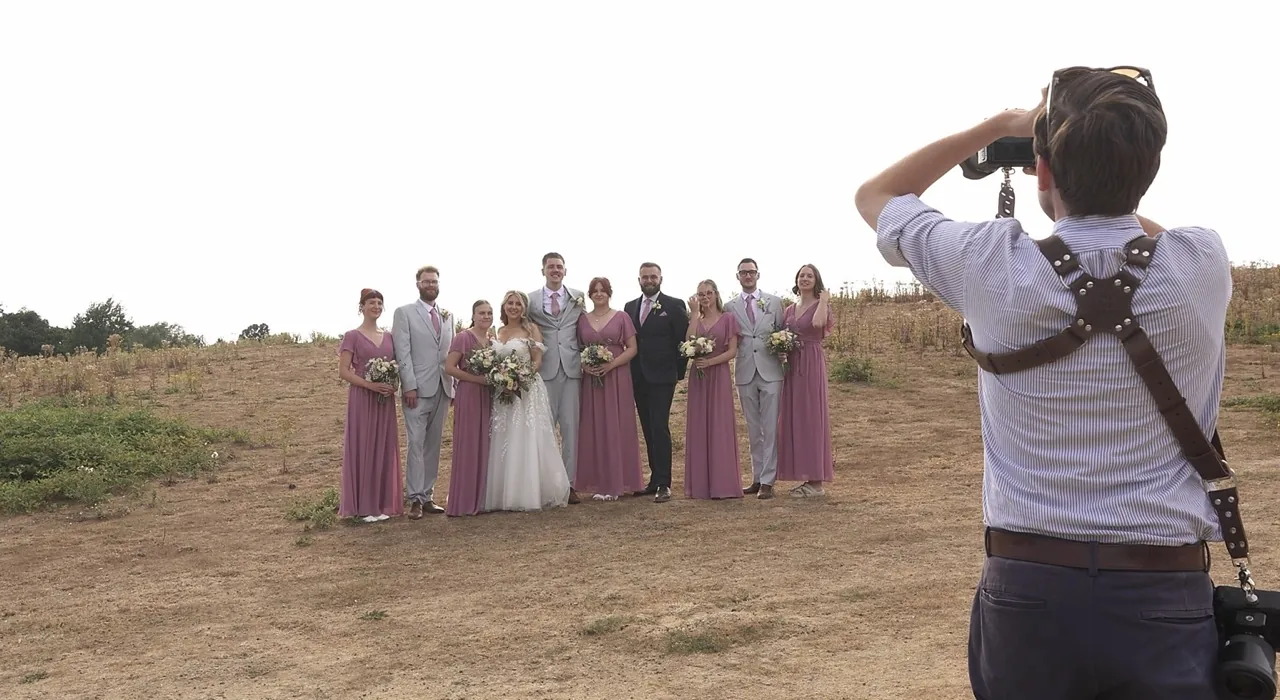 A photographer lining up a group portrait of the wedding party in an open field